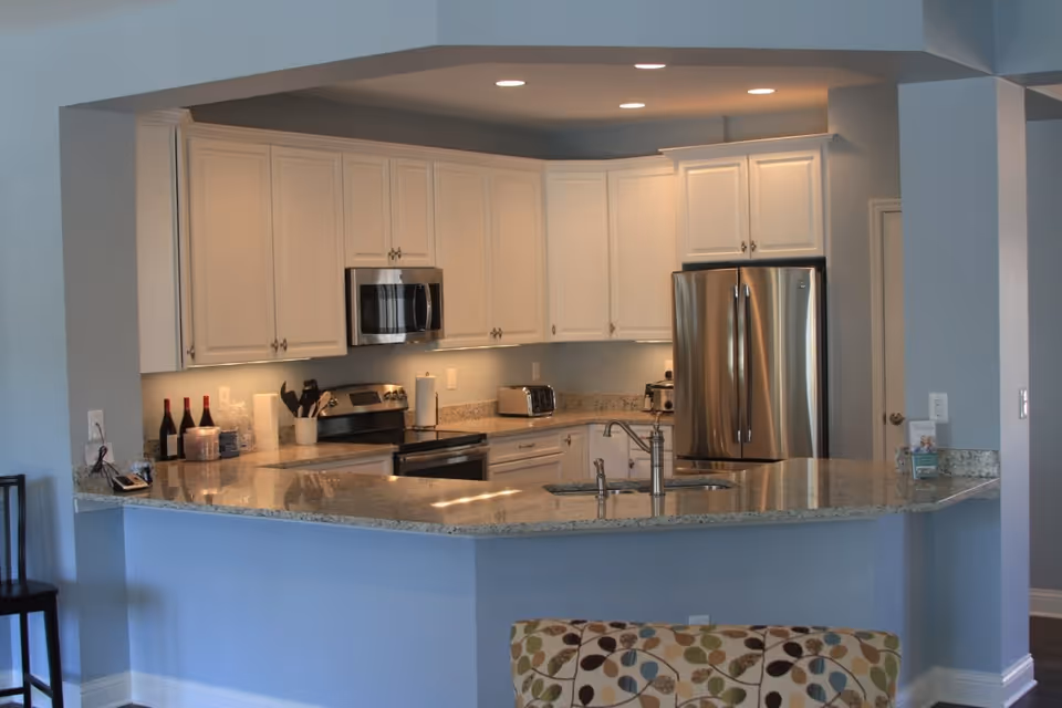 Modern kitchen with white cabinets, stainless steel refrigerator, microwave, and stove. The kitchen features a granite countertop with a built-in sink and faucet. There are various kitchen items on the counter including wine bottles, a toaster, and paper towels. The walls are painted light blue and there is a patterned chair partially visible in the foreground.