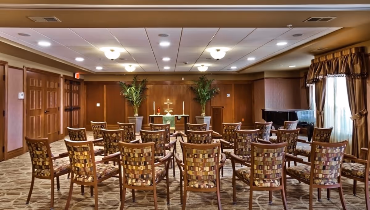 A small chapel or worship room with rows of patterned chairs facing an altar with candles, a cross, and two large potted plants. The room has wood-paneled walls, a carpeted floor, ceiling lights, and a grand piano near a window with draped curtains.