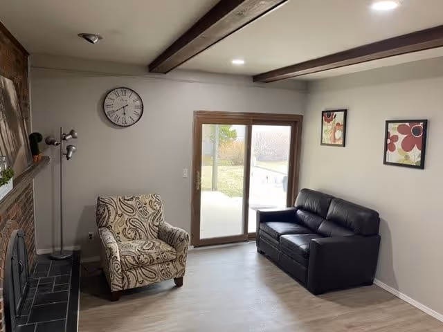 A cozy living room with a patterned armchair and a black leather sofa facing a brick fireplace. There is a floor lamp next to the armchair, a wall clock above it, and two framed floral artworks on the wall above the sofa. A sliding glass door leads to an outdoor area with grass and trees visible.