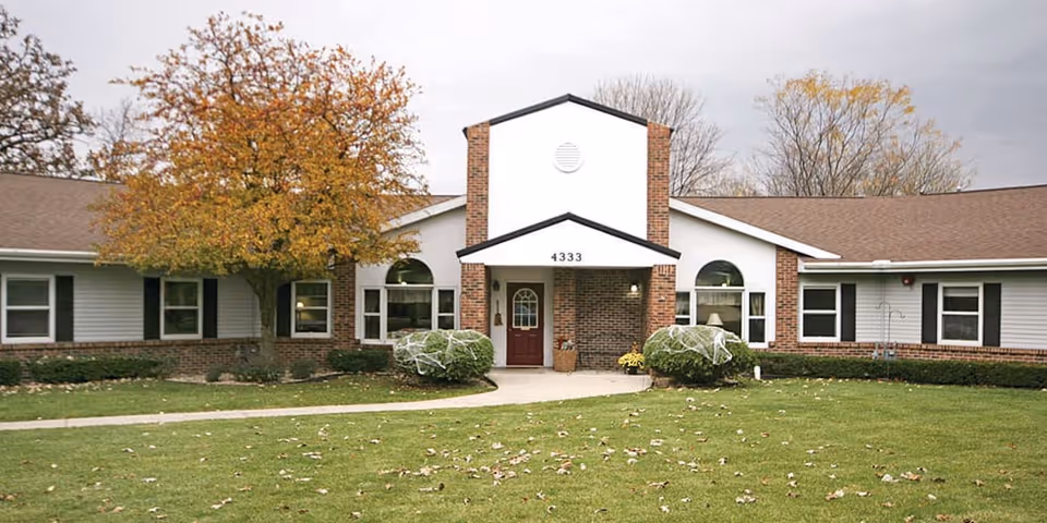 Exterior front view of a single-story senior living facility building with a brown roof, white siding, and brick accents. There is a central entrance with a red door under a small covered porch. The building is surrounded by a green lawn with scattered fallen leaves and a few trees with autumn-colored foliage.