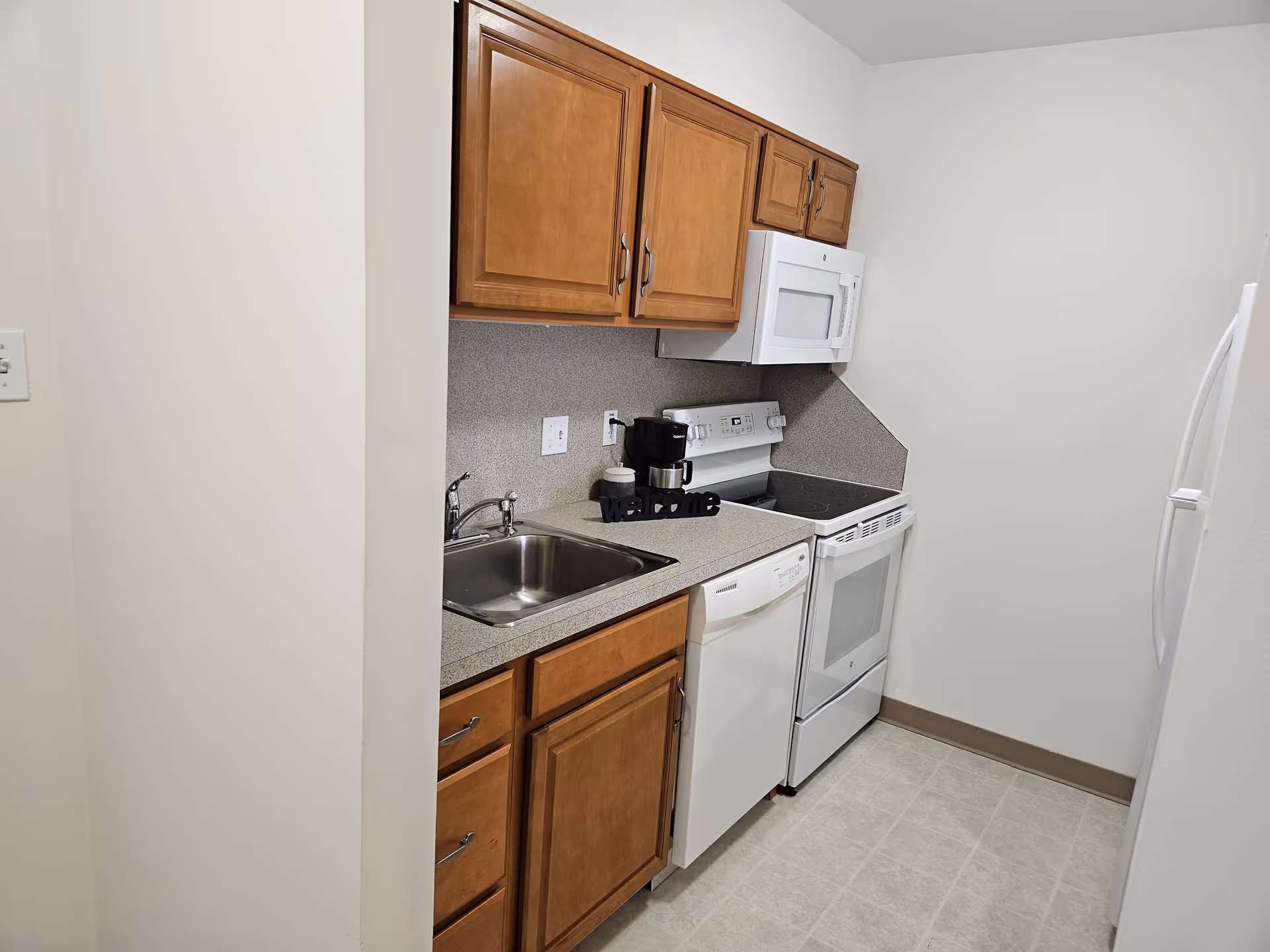 A small kitchen area with wooden cabinets, a stainless steel sink, a white dishwasher, a white electric stove with an oven, a white microwave mounted above the stove, and a white refrigerator. There is a coffee maker and a decorative 'welcome' sign on the countertop.