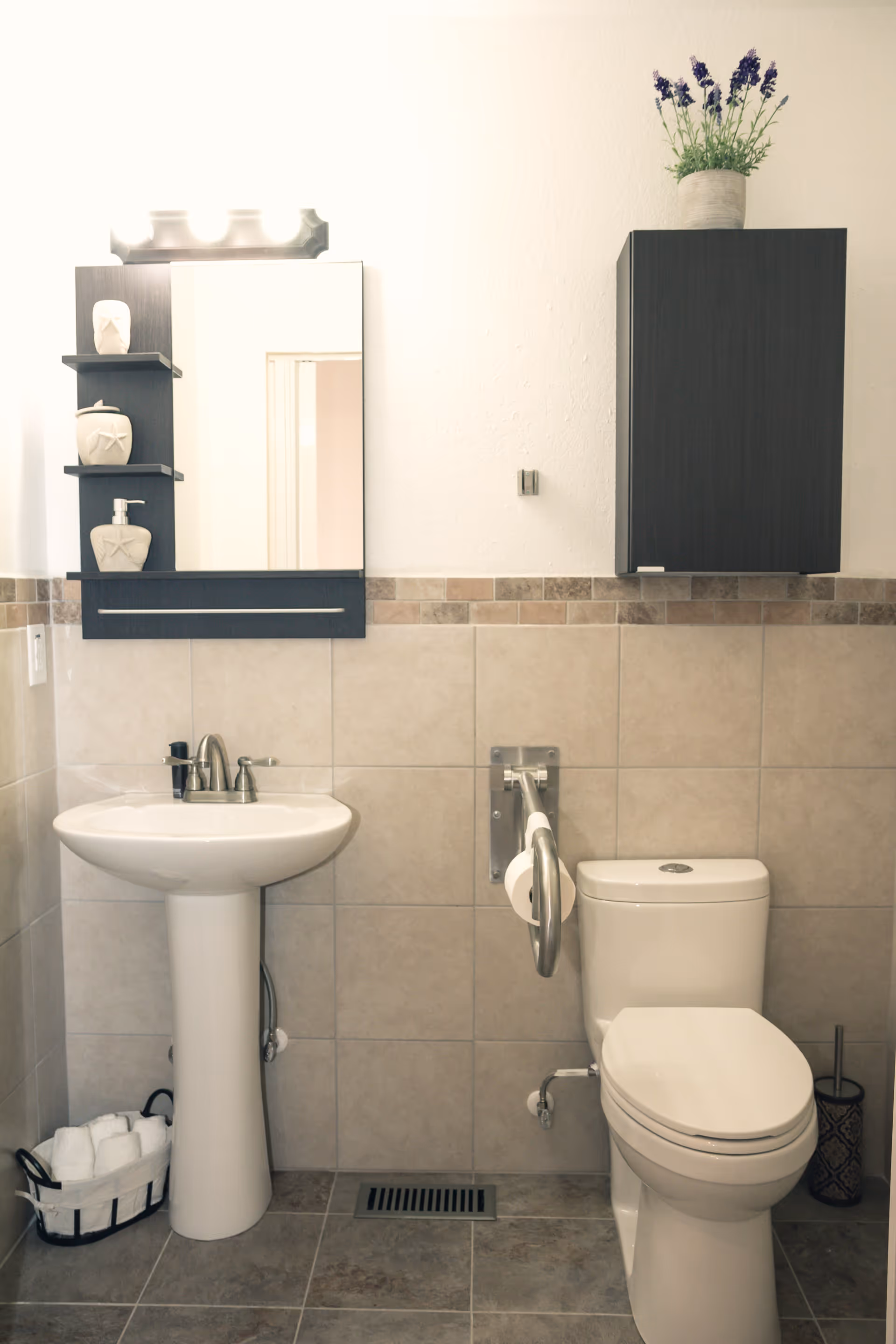 A clean bathroom with beige tiled walls and floor. It features a white pedestal sink with a silver faucet, a rectangular mirror with shelves on the left holding decorative items, and a dark wood cabinet mounted on the wall with a potted plant on top. There is a white toilet with a metal grab bar and toilet paper holder attached to the wall beside it. A basket with rolled white towels is placed on the floor near the sink.
