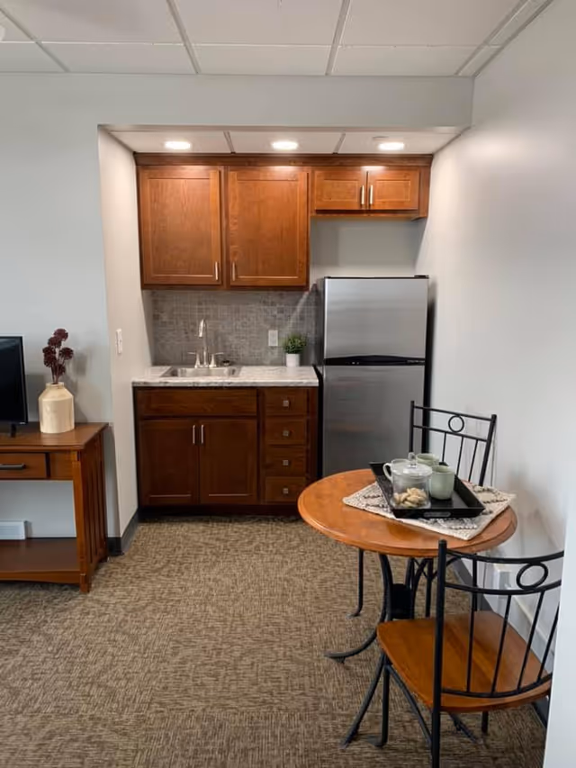 Small kitchenette area with wooden cabinets, a stainless steel mini refrigerator, a sink, and a tiled backsplash. Next to the kitchenette is a round wooden table with two metal chairs, set with a tray holding a teapot, cups, and some snacks. A wooden side table with a vase and decorative flowers is visible on the left side.