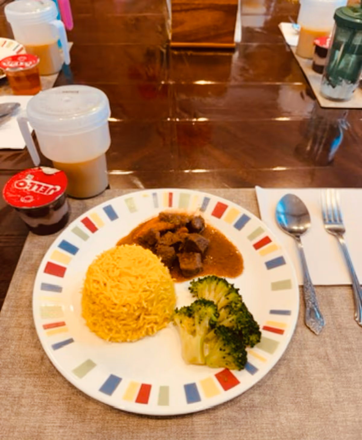 A plate of food on a table with yellow rice, beef stew, and steamed broccoli. Next to the plate are a spoon and fork on a napkin. There is a cup with a lid containing a beverage and a small container of Jell-O on the left side of the plate. The table has a brown tablecloth and other similar place settings are visible in the background.