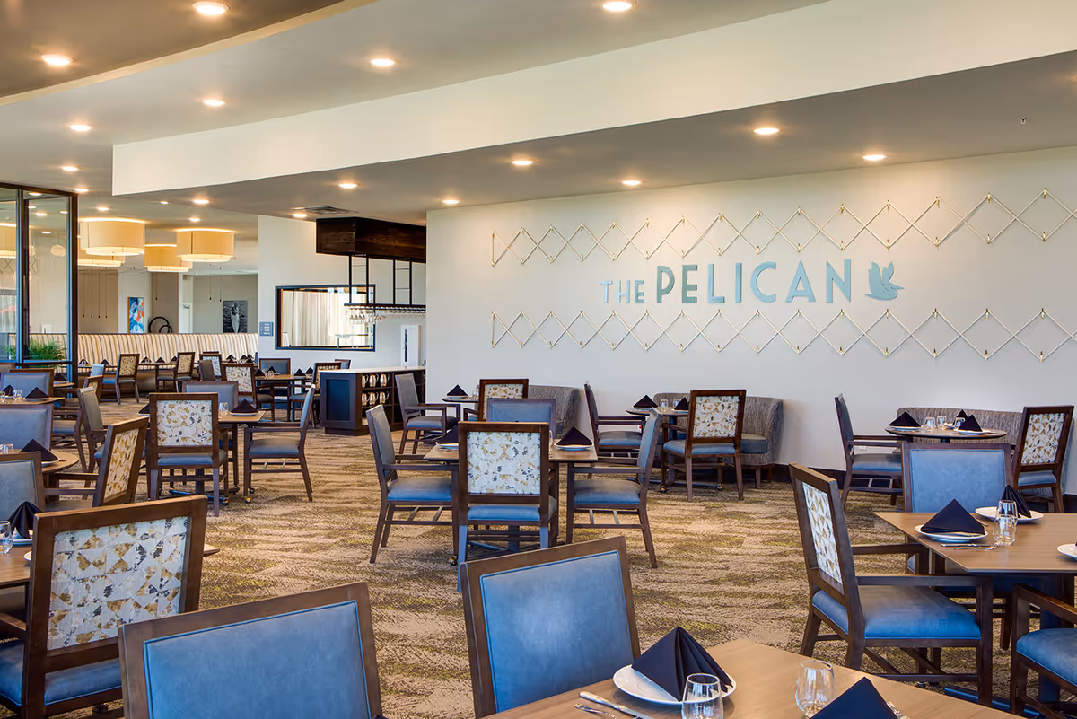 A spacious dining room with multiple wooden tables and chairs, each table set with plates, glasses, silverware, and black folded napkins. The wall features a decorative design with the words 'THE PELICAN' and a pelican logo. The room is well-lit with ceiling lights and large hanging lamps in the background.
