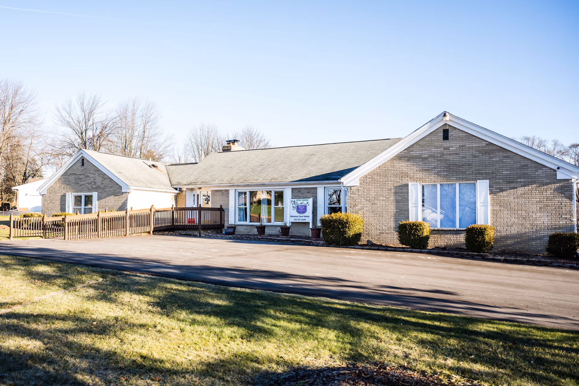 Exterior view of a single-story brick building with white trim and several windows, surrounded by a paved driveway and some bushes. The building has a sign near the entrance that reads 'Memory Lane Assisted Living (Stoneham House)'. Trees without leaves are visible in the background under a clear blue sky.