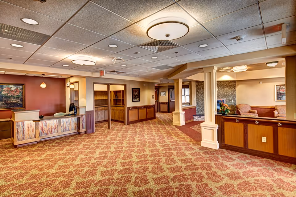 Interior view of a senior living facility lobby area with patterned carpet, a reception desk on the left, wooden cabinetry, and a seating area with armchairs on the right. The ceiling has recessed lighting and round ceiling fixtures, and the walls are decorated with framed artwork.