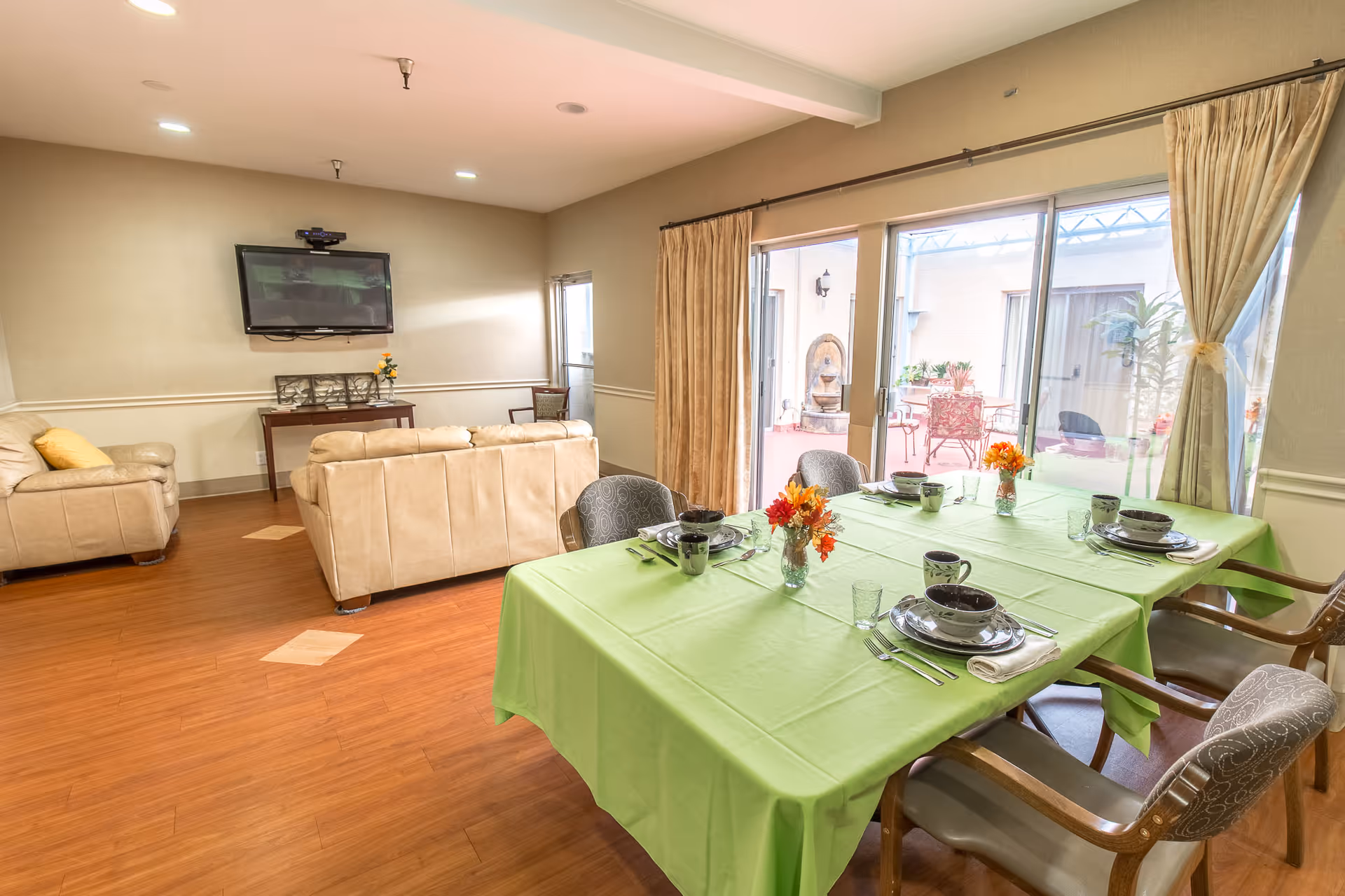 Bright common room with a green-clothed dining table set for four, beige sofas facing a wall-mounted TV, and sliding glass doors to a patio.