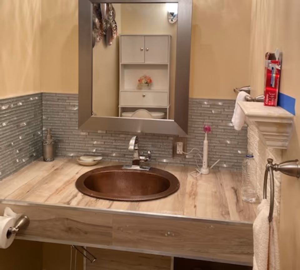 Bathroom sink area with a round copper basin set in a wood-look countertop, mirror, toothbrush and towels.