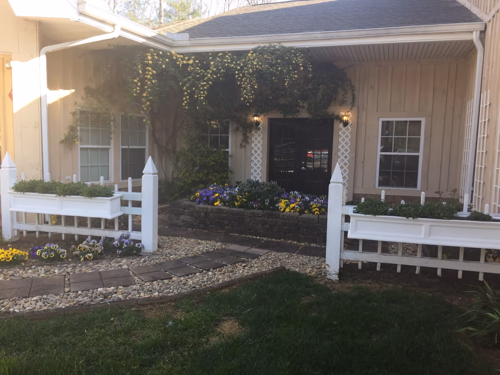 Outdoor view of a building entrance with beige siding, two windows, and a black double door. The entrance is decorated with hanging yellow flowers, a flower bed with purple and yellow flowers, white wooden fences with planter boxes, and a stone pathway surrounded by grass and pebbles.