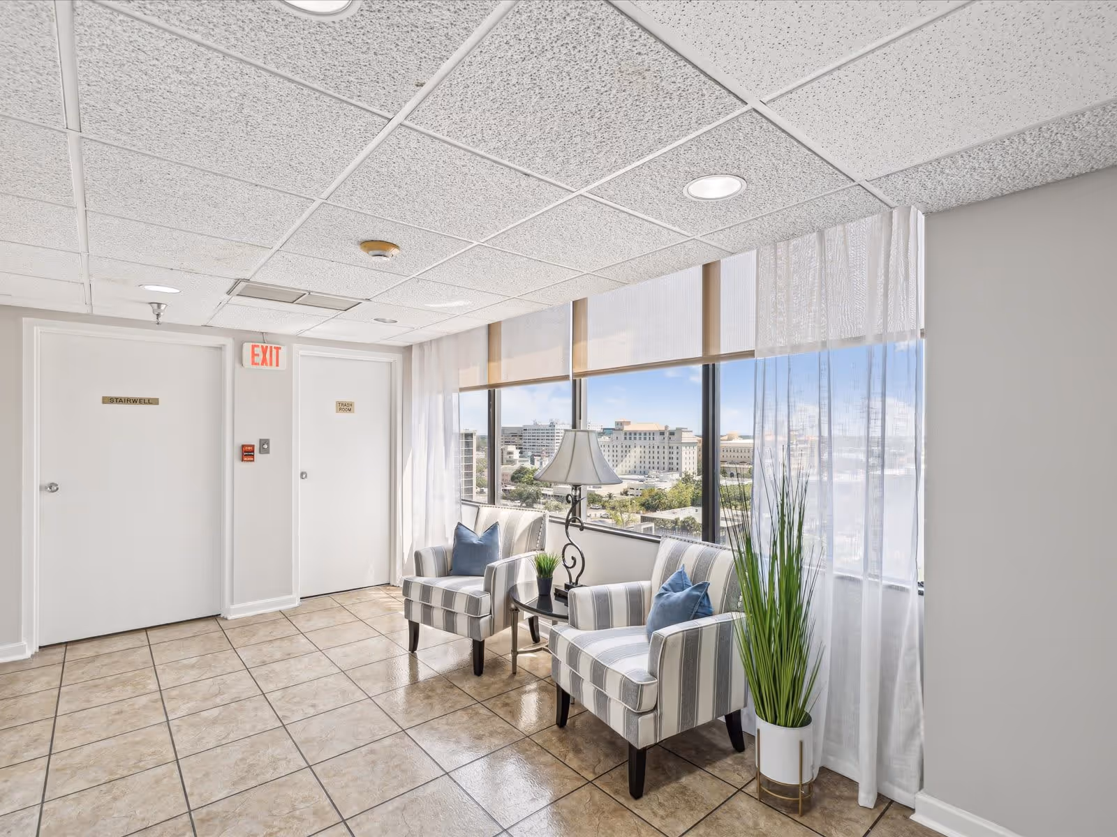 A bright interior corner seating area with two striped armchairs, each with a blue pillow, a small round table with a lamp and a small plant, large windows with sheer white curtains, and a view of city buildings outside. There are two white doors labeled 'Stairwell' and 'Trash Room' with an exit sign above.