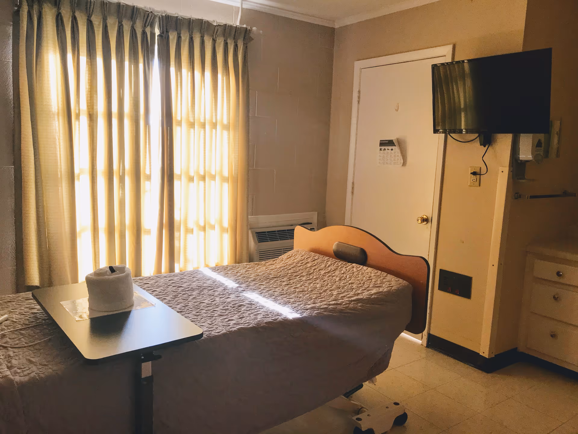 A nursing facility bedroom with a single bed covered in a beige quilt. A small overbed table with a rolled towel is positioned on the bed. Beige curtains cover a window letting in sunlight. A flat-screen TV is mounted on the wall next to a closed door. There is a small air conditioning unit below the window and a cabinet with drawers on the right side.