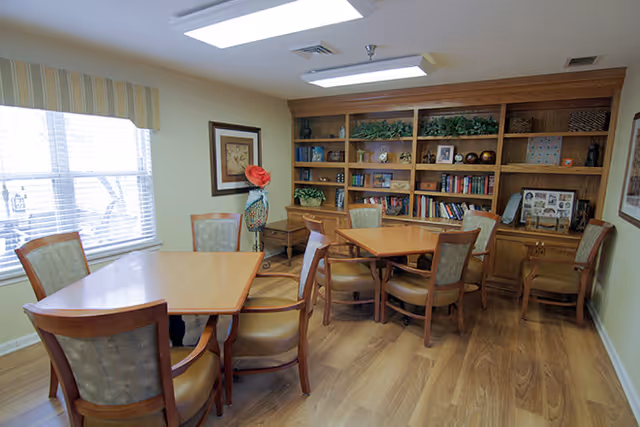 Communal activity room with wooden tables and chairs, built-in bookshelves, and a large window.