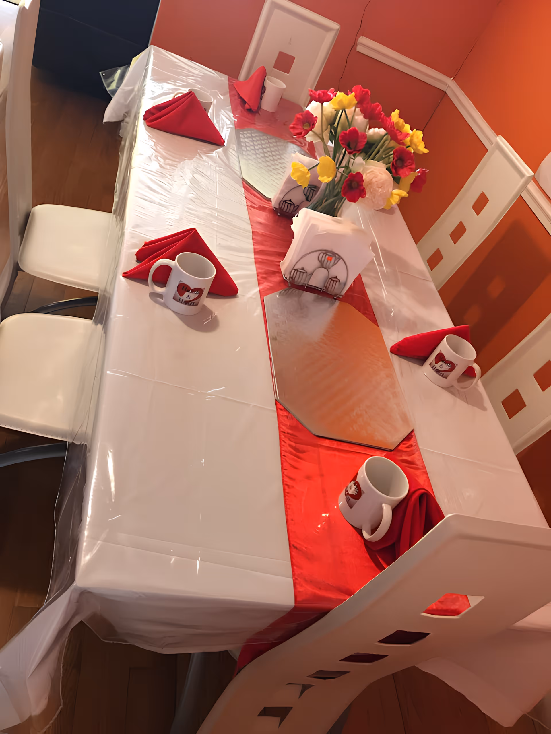 Dining table set with a red table runner, folded red napkins, white mugs and a vase of flowers in an orange-walled room.