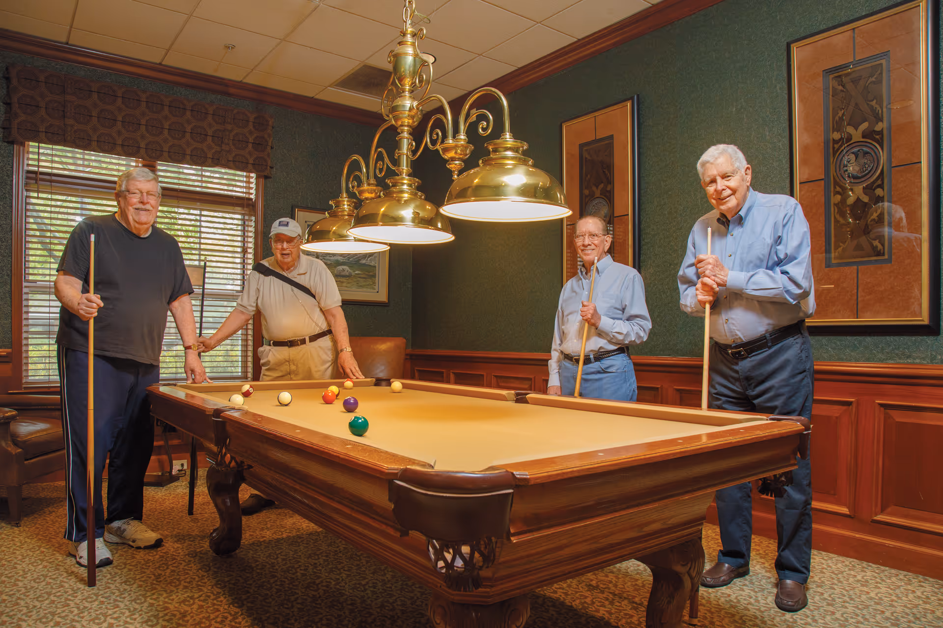 Four elderly men standing around a billiards table in a room with green walls, wooden paneling, and framed artwork. The men are holding pool cues and smiling. There is a window with blinds and a valance, and a brass light fixture hanging above the pool table.