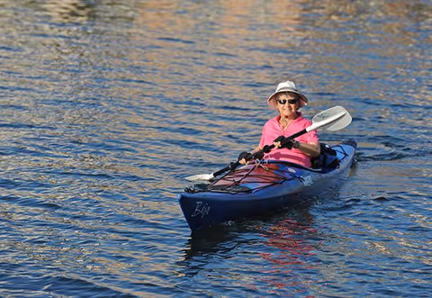 An elderly person wearing a white hat, sunglasses, and a pink shirt paddling a blue kayak on calm water.