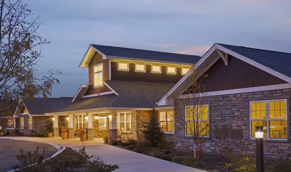 Exterior view of a single-story building with stone and wood siding, illuminated windows, and a paved walkway leading to the entrance during dusk.