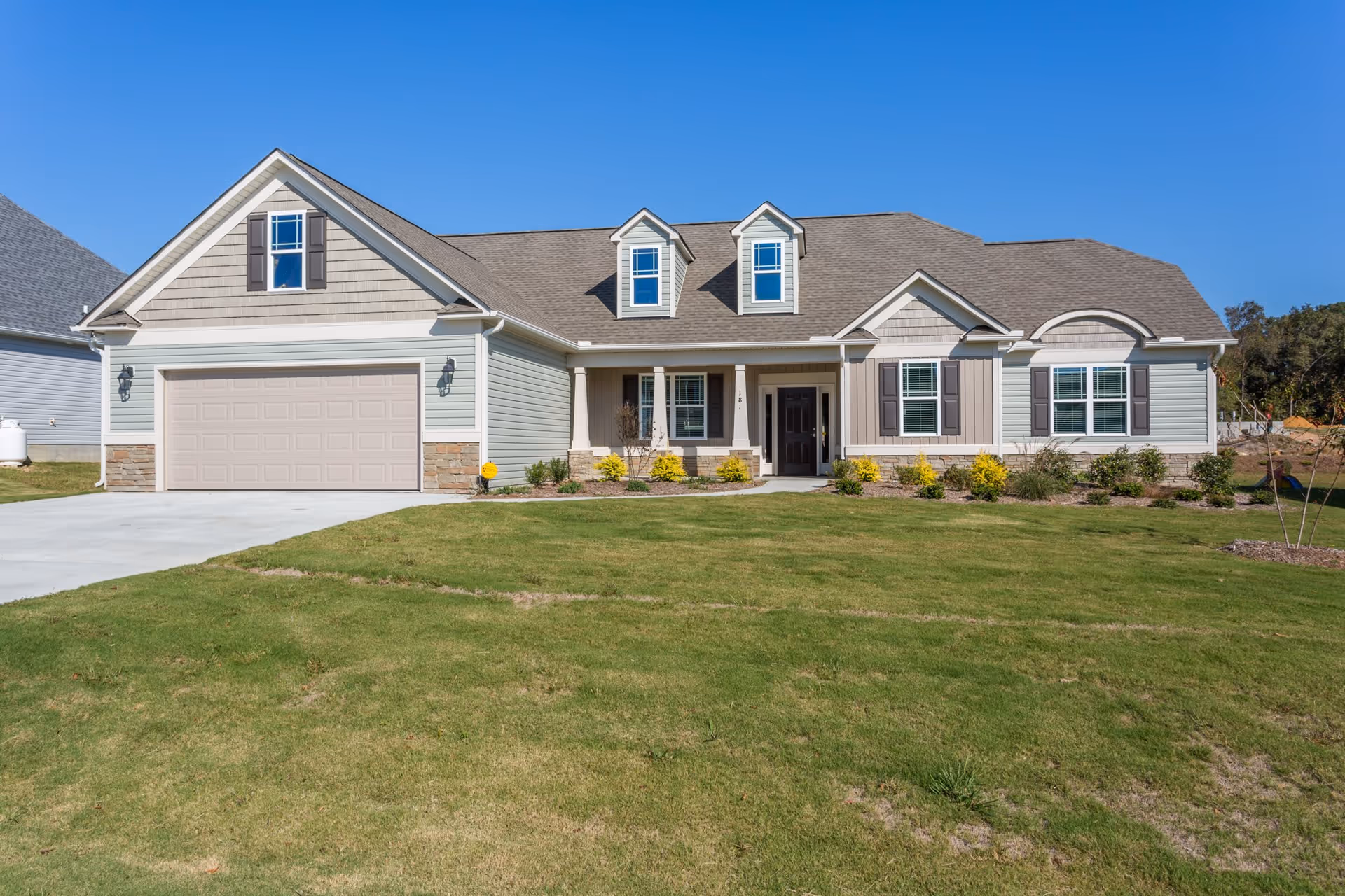 Front exterior view of a single-story house with a two-car garage, beige siding, stone accents, and a well-maintained lawn under a clear blue sky.