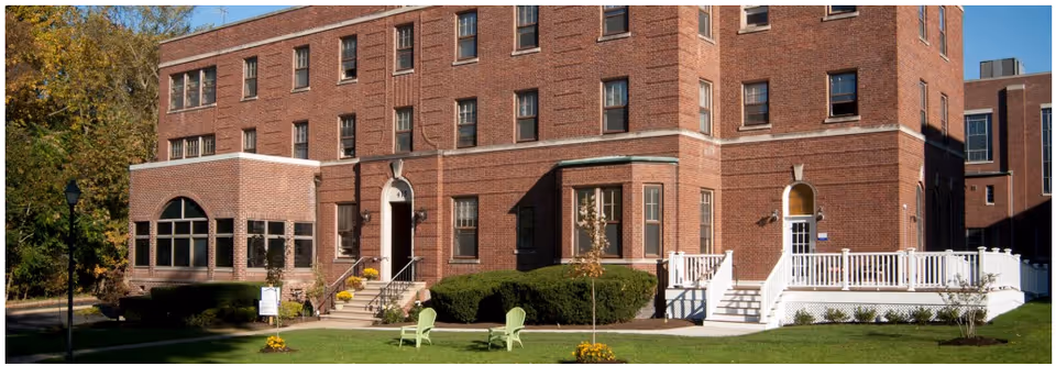 Exterior view of a large brick senior living facility building with multiple windows, two entrances with stairs, a white railing on one side, green lawn with two green chairs, small trees, and bushes under a clear blue sky.