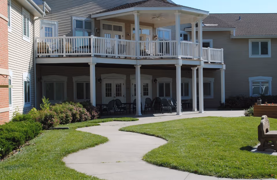 Outdoor courtyard of a senior living facility featuring a two-story covered patio with chairs and a winding concrete walkway through a lawn.
