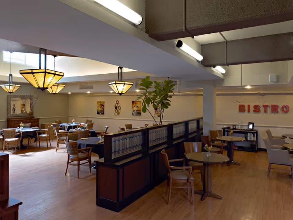 Interior view of a dining area in a senior living facility with several tables and chairs arranged neatly. The room features warm lighting with decorative hanging lamps, a wooden floor, and a partition with decorative tiles. On the far wall, there is a sign that reads 'BISTRO' in large red letters. The space is cozy and inviting, designed for communal dining and socializing.