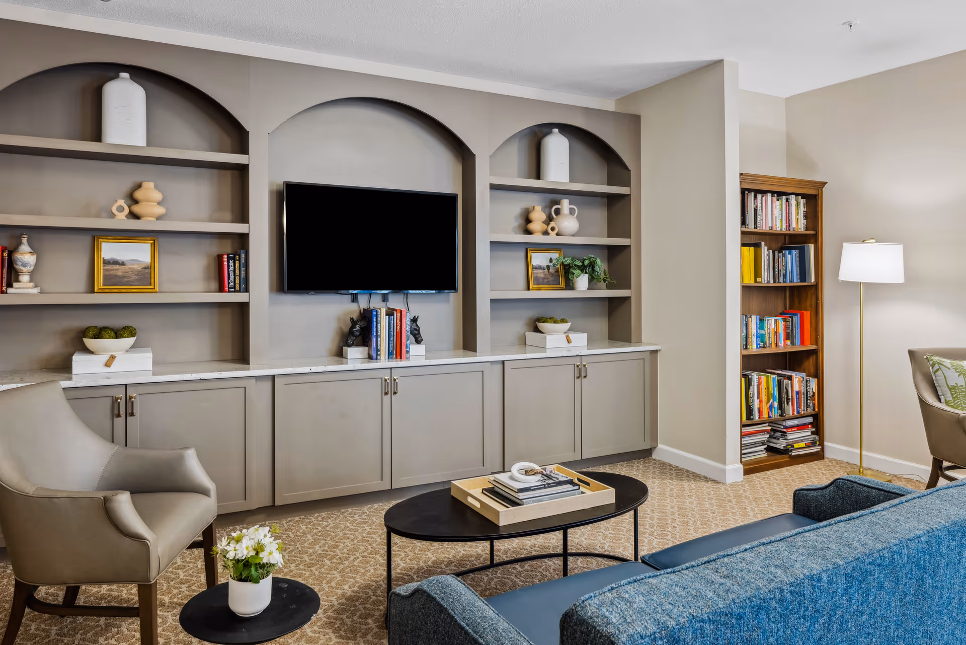 A cozy living room with built-in shelves and cabinets painted in gray. The shelves hold decorative items, books, and framed pictures. A flat-screen TV is mounted in the center of the shelving unit. The room features a blue sofa, a beige armchair, a small round black side table with a white flower pot, a black oval coffee table with books and a tray, a wooden bookshelf filled with books, and a floor lamp with a white shade. The carpet has a subtle patterned design.