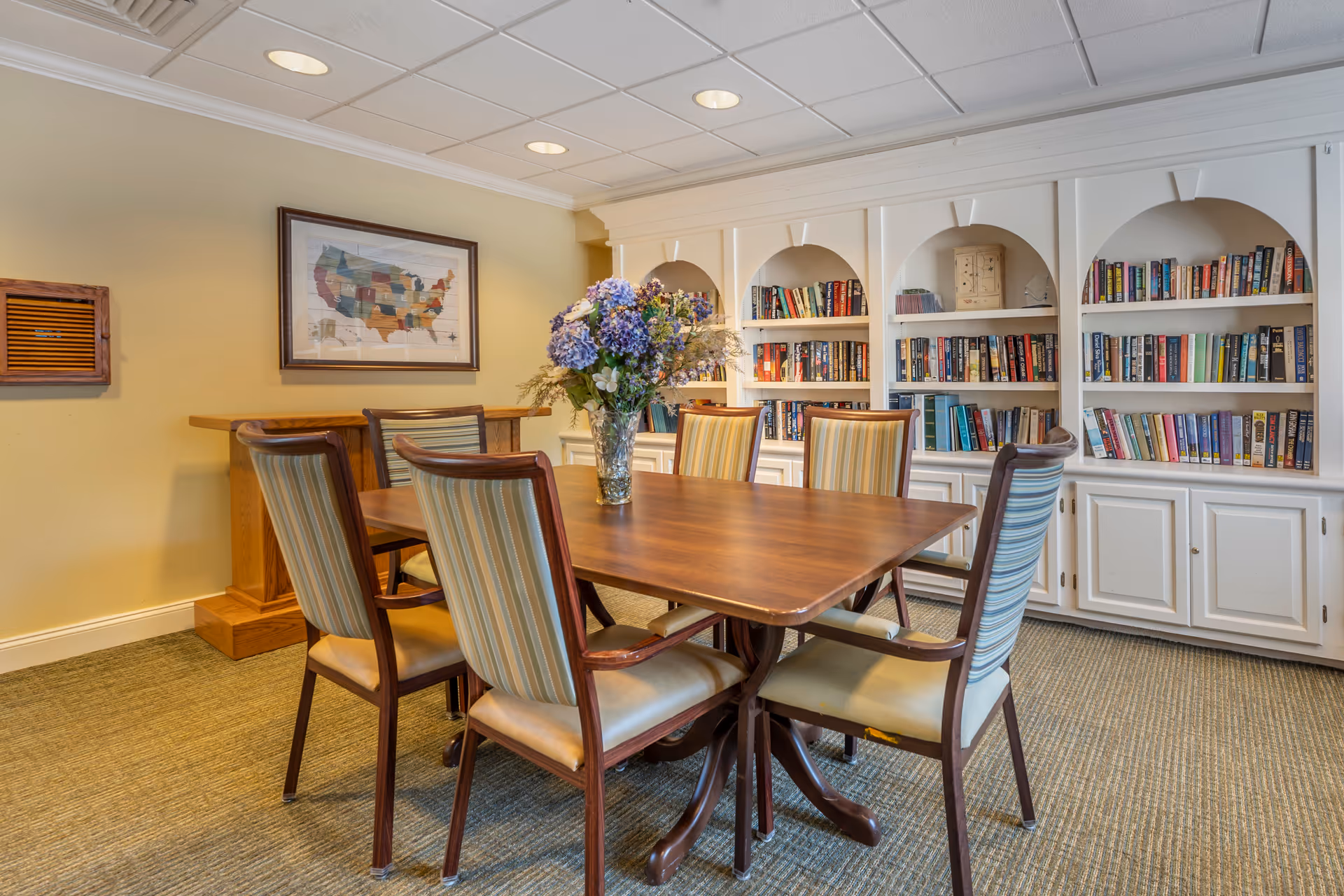 A cozy room with a wooden table surrounded by six striped cushioned chairs. A vase with purple and white flowers is placed on the table. Behind the table, there is a white built-in bookshelf filled with books. A framed map of the United States hangs on the beige wall to the left.
