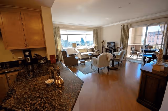 A bright and spacious living room area with wooden flooring, featuring a seating arrangement of armchairs and a sofa around a coffee table. Large windows and a sliding glass door lead to a balcony with outdoor furniture. The foreground shows a kitchen counter with granite countertops and wooden cabinets.