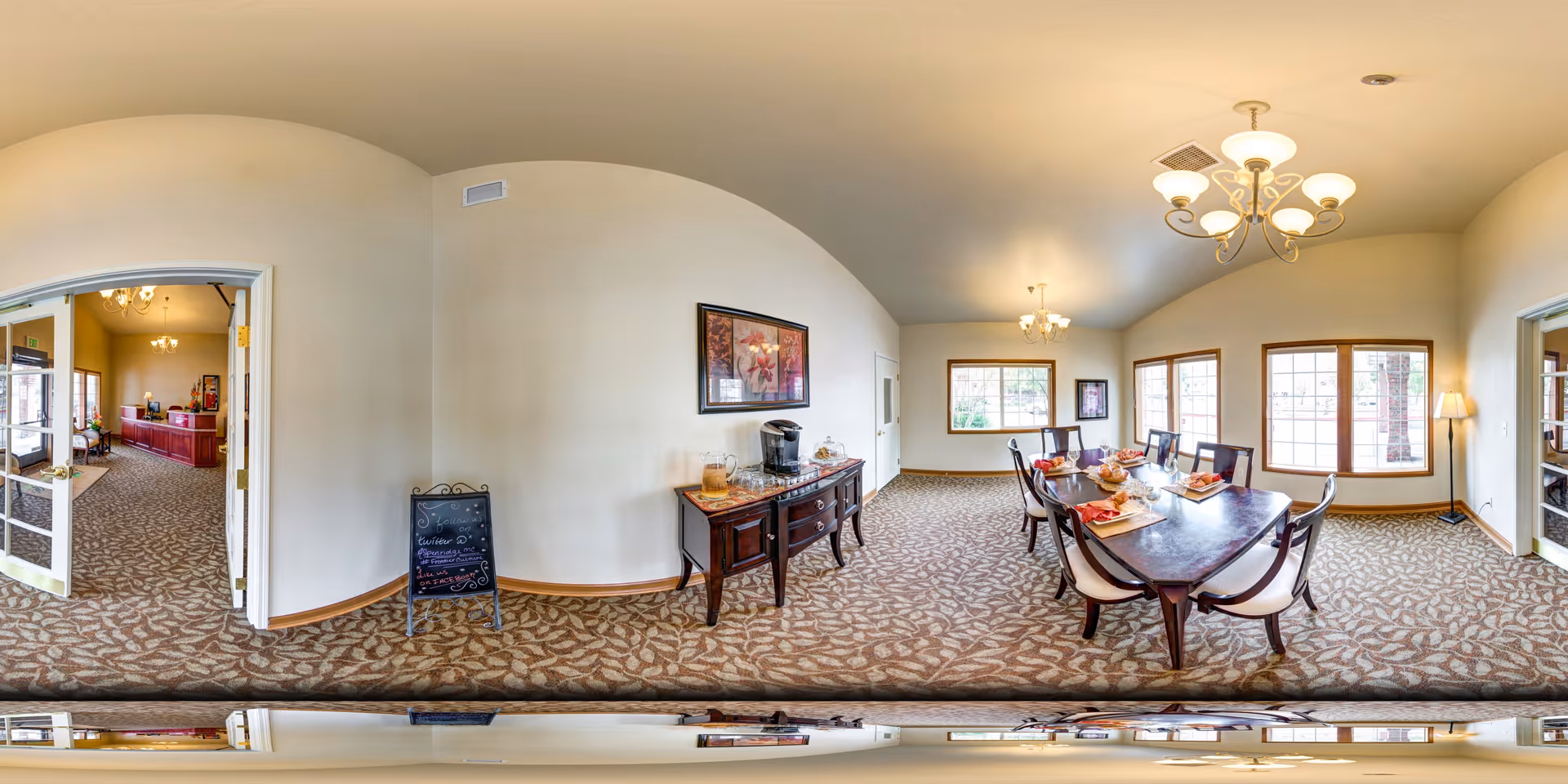 A spacious dining room in Aspen Ridge Memory Care with a large wooden dining table set for a meal, surrounded by six chairs. The room features beige walls, patterned carpet, two chandeliers, and large windows letting in natural light. A sideboard with a coffee maker and decorative items is against one wall, and a chalkboard sign is placed near the entrance. Adjacent rooms are visible through open double doors.