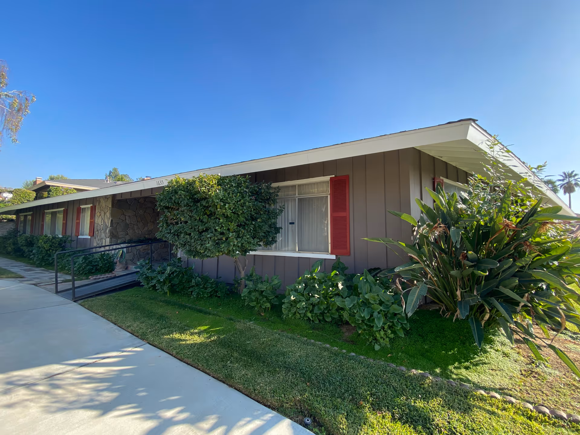 Single-story mid-century building with brown siding, red shutters, a wheelchair ramp and green landscaping under a clear blue sky.