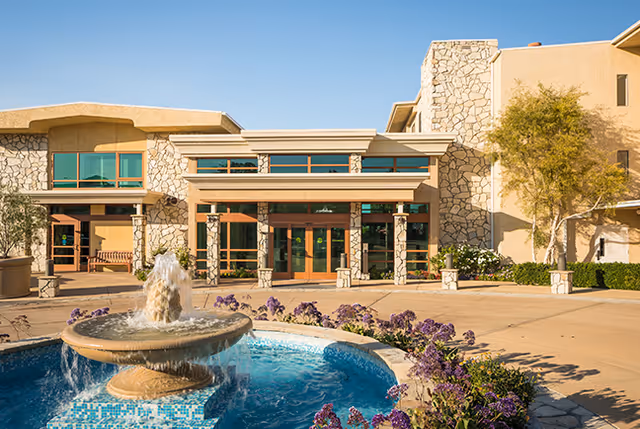 A fountain and purple flowerbeds in a courtyard leading to the stone-faced entrance of a senior living building under a clear blue sky.