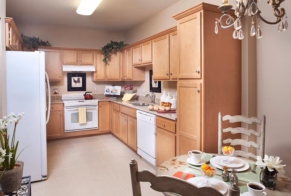 A bright kitchen with light wood cabinets, a white refrigerator, stove, and dishwasher. There is a small round dining table set for two with plates, coffee cups, and fruit parfaits. Decorative plants and kitchen accessories are placed on the countertops and above the cabinets.