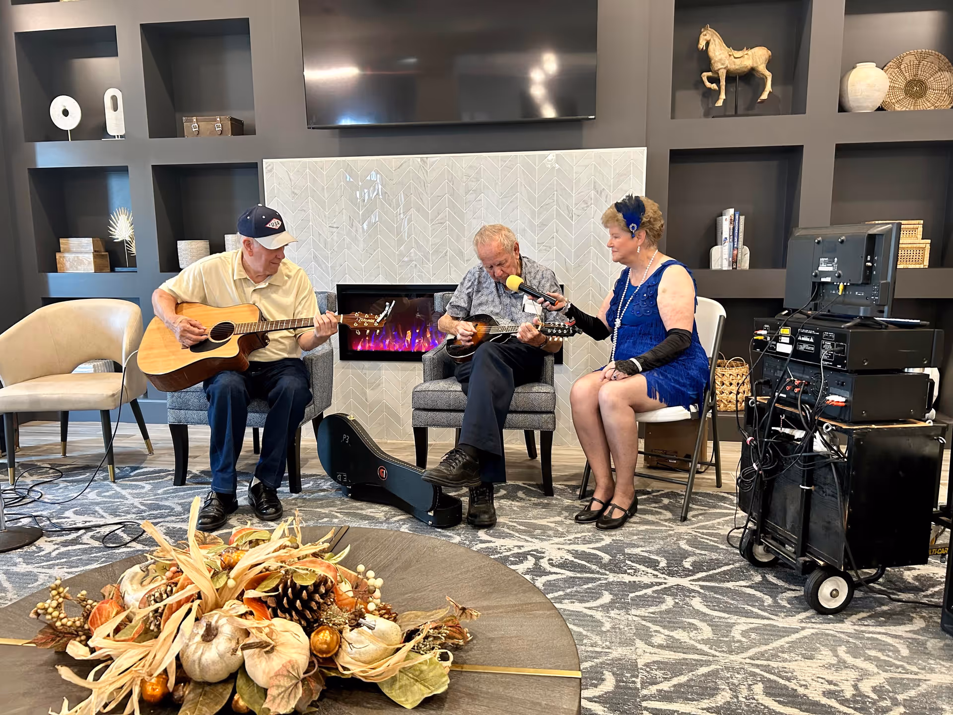 Three elderly individuals in a cozy living room setting; two men are playing string instruments while a woman in a blue dress holds a microphone to one of the men. They are seated in front of a modern electric fireplace with built-in shelves displaying decorative items. A round table with a fall-themed centerpiece is in the foreground.