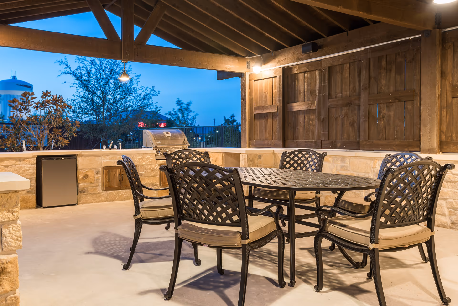 Covered outdoor patio area with a metal table and six cushioned chairs. The space includes a built-in stone countertop with a small refrigerator and a stainless steel grill. Wooden beams and fencing enclose the area, with trees and a water tower visible in the background during twilight.