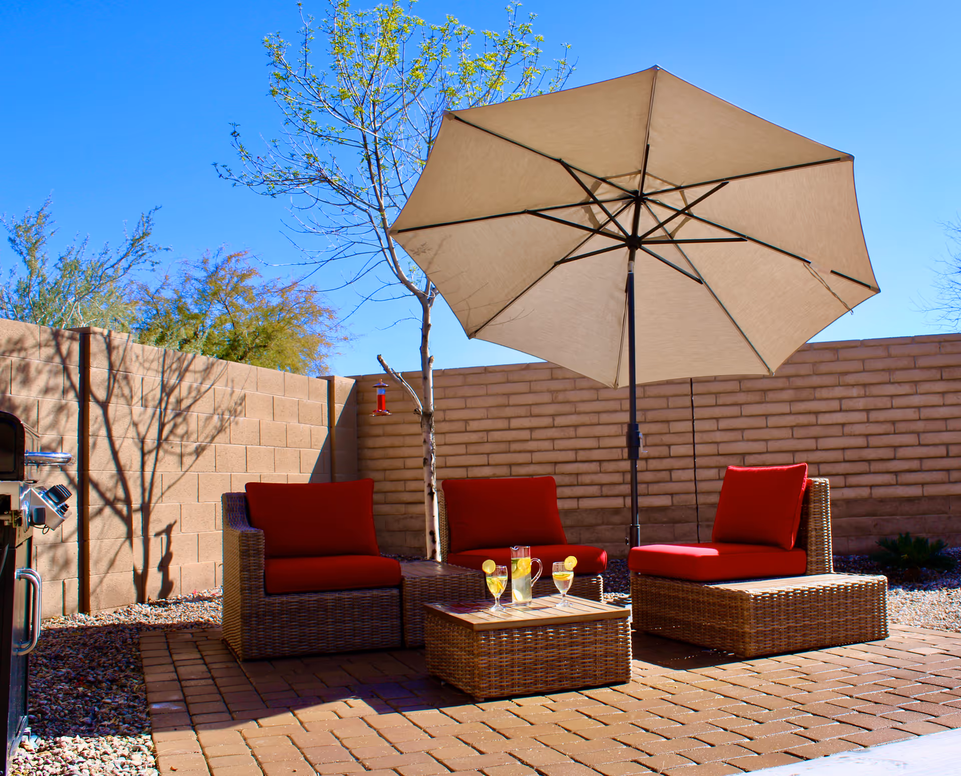Outdoor patio area with three wicker chairs featuring red cushions, a matching wicker coffee table with a pitcher of lemonade and three glasses, a large beige umbrella providing shade, a brick wall surrounding the space, a small tree, and a barbecue grill on the left side.