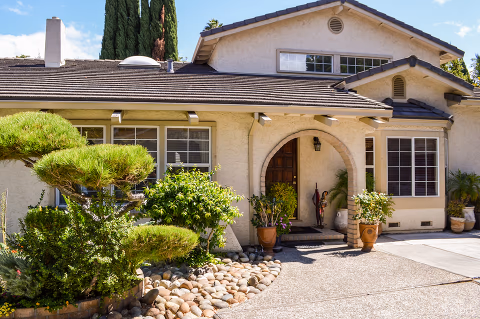Front exterior view of a beige stucco house with a tiled roof, an arched entrance with a wooden door, potted plants, and manicured bushes and trees in the front yard under a blue sky.