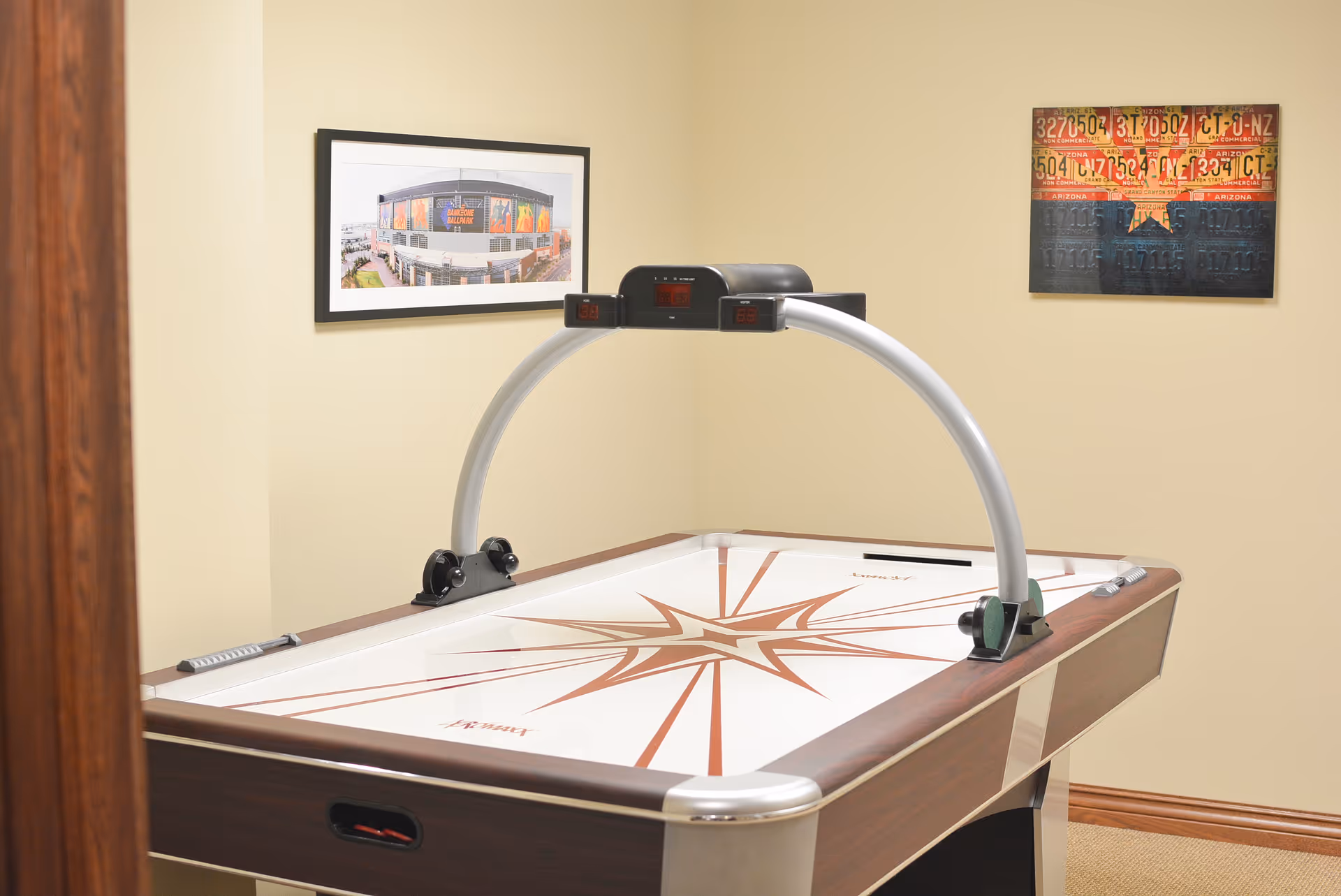 A room with an air hockey table featuring a star design on its surface. The table has a digital scoreboard arching over it. The walls are beige, adorned with two framed pictures: one showing a building and the other a colorful artwork with numbers and a star. The room has wooden trim around the door and baseboards.
