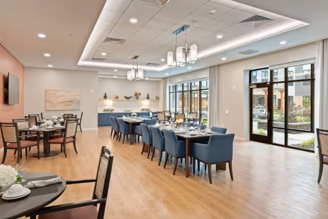 A spacious and well-lit dining room with wooden flooring, multiple dining tables set with white plates, cups, and napkins. The room features blue upholstered chairs around a long table and other chairs with woven backs around smaller tables. Large windows and glass doors allow natural light to enter, and there are modern light fixtures hanging from the ceiling. A sideboard with decorative items and a painting on the wall complete the room.