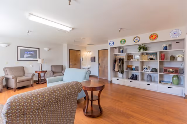 A spacious and well-lit common area with wooden flooring, featuring several upholstered chairs and a small round wooden table. On the right side, there is a large white shelving unit displaying various decorative items, clothing, and military insignia plaques above it. Two wooden doors and a wall-mounted sink with a mirror are visible in the background.