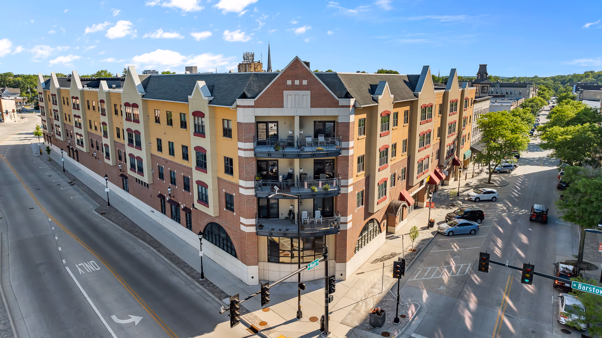A large multi-story brick and beige building with balconies on the corner, situated at an intersection with traffic lights and parked cars along the streets. The sky is blue with scattered clouds and there are trees lining the streets.