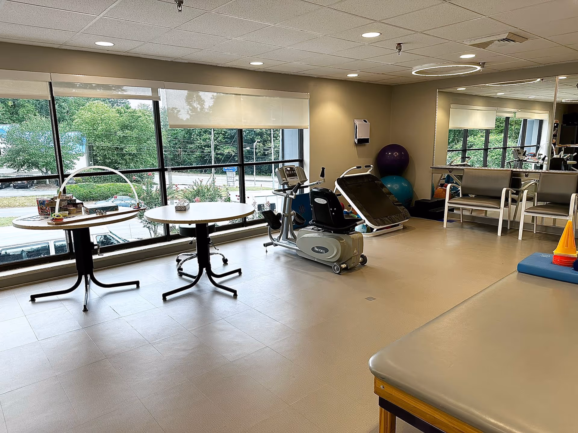 A bright rehabilitation or physical therapy room with large windows showing greenery outside. The room contains exercise equipment including a recumbent bike, therapy balls, a padded therapy table, two round tables with items on them, chairs, and a large mirror on one wall.