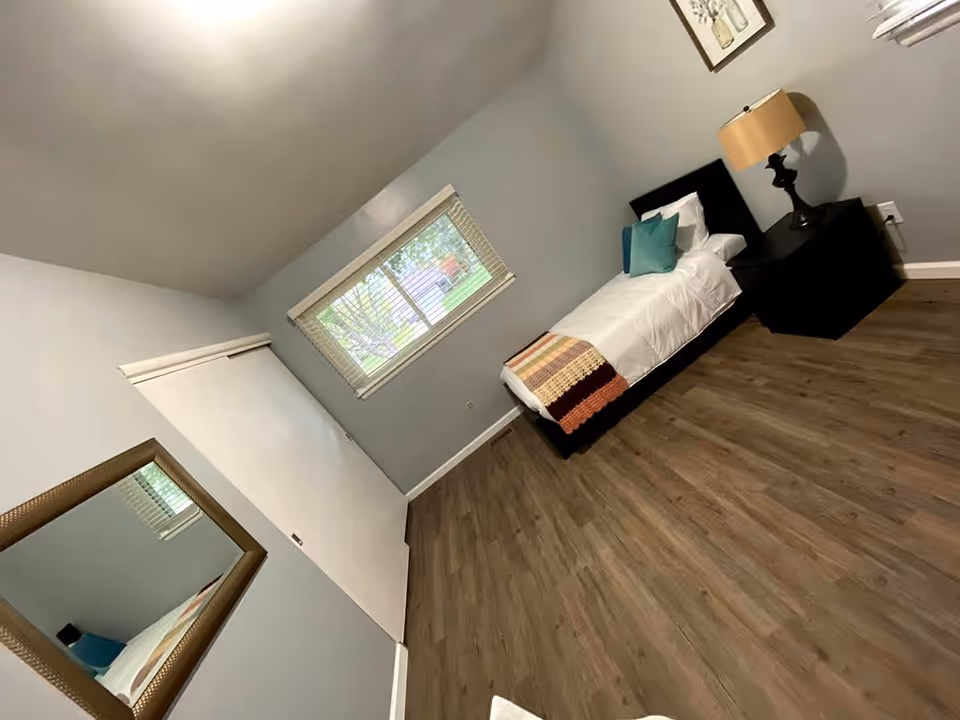 A simple bedroom with a single bed against the wall, a nightstand with a lamp, a window with blinds, a framed picture on the wall, a large mirror on another wall, and wood flooring.