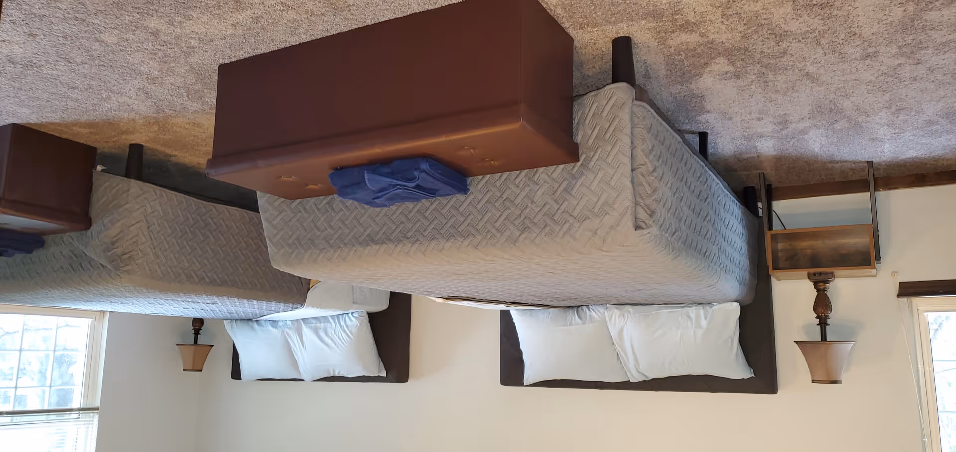 Two twin beds with white pillows and foot ottomans in a simple, carpeted bedroom with bedside lamps and a window.