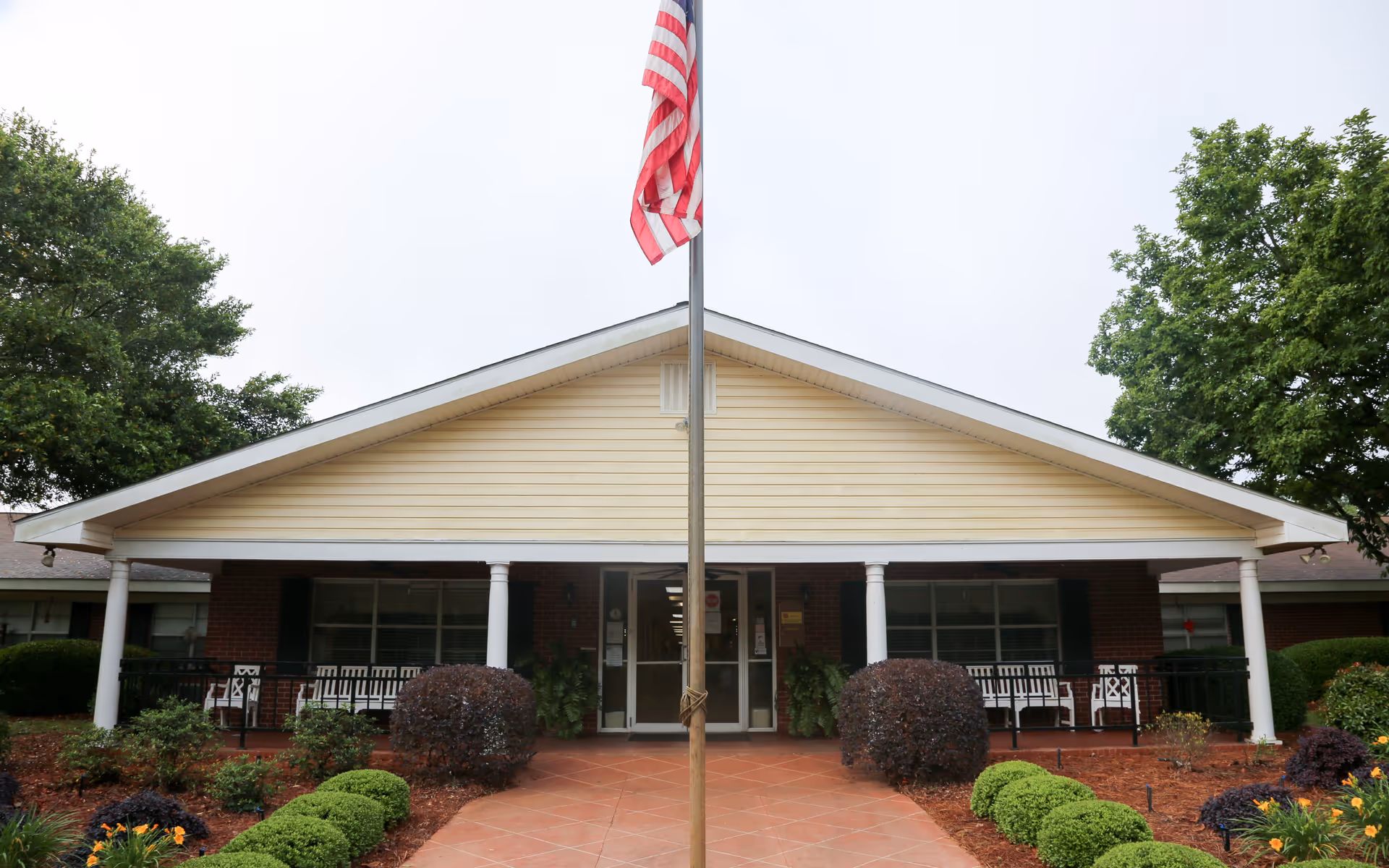 Front exterior view of a single-story building with a peaked roof, white columns, and a brick facade. There is a flagpole with an American flag in the center of a paved walkway leading to the entrance. The entrance has glass double doors, and there are bushes and landscaped plants on either side of the walkway. Trees are visible on both sides of the building.