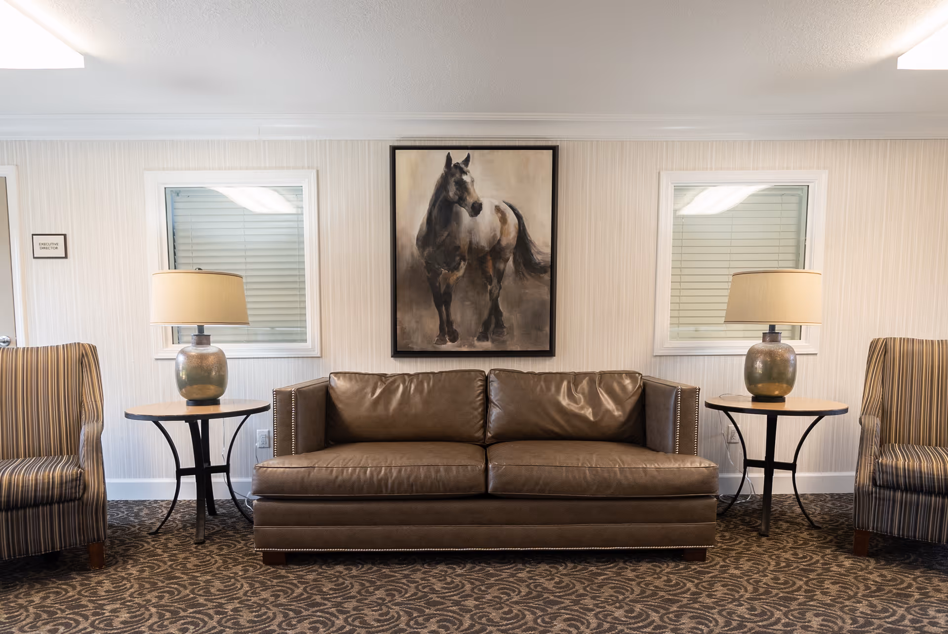 A cozy seating area in a memory care community featuring a brown leather sofa centered between two round wooden side tables with matching lamps. On either side of the sofa are striped armchairs. Above the sofa hangs a framed painting of a horse. The walls are light-colored with two small windows behind the sofa.