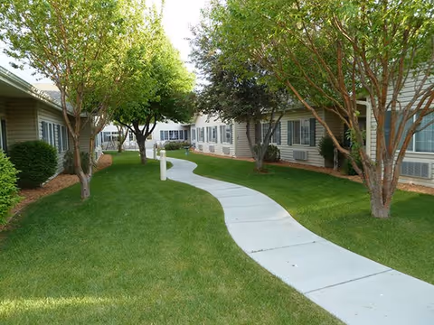 Curving concrete walkway through a grassy courtyard lined with trees between single-story assisted living buildings.