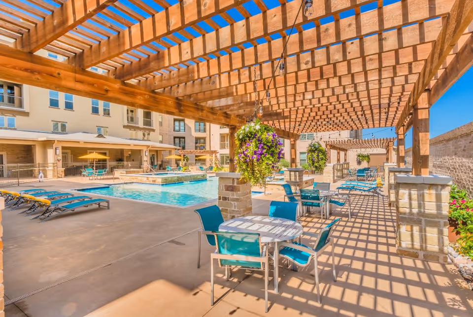 Outdoor pool and lounge area with a wooden pergola, hanging plants, tables and turquoise lounge chairs beside a multi-story building.