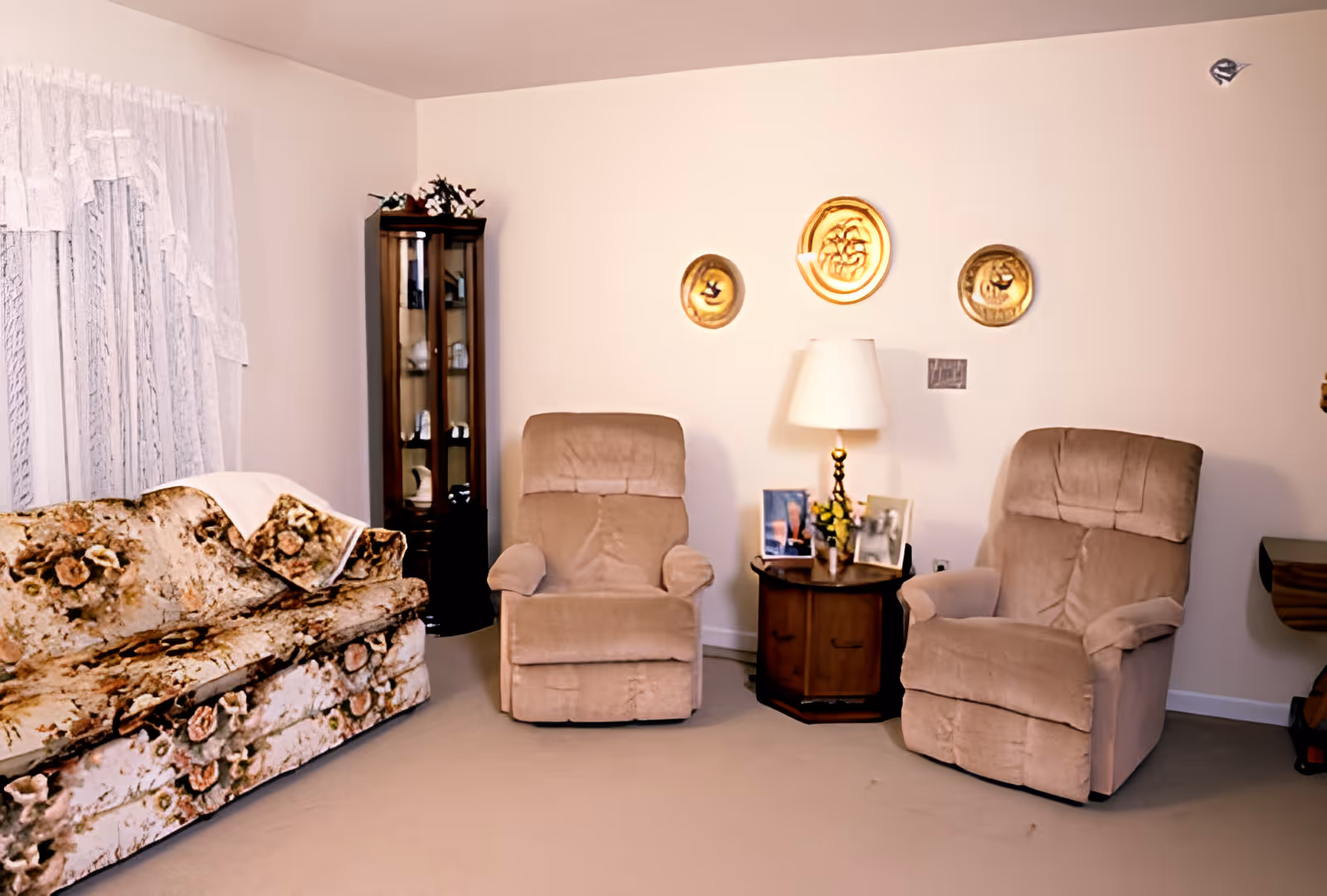 A cozy living room with a floral sofa, two beige recliners, an end table with a lamp and photos, and decorative plates on the wall.