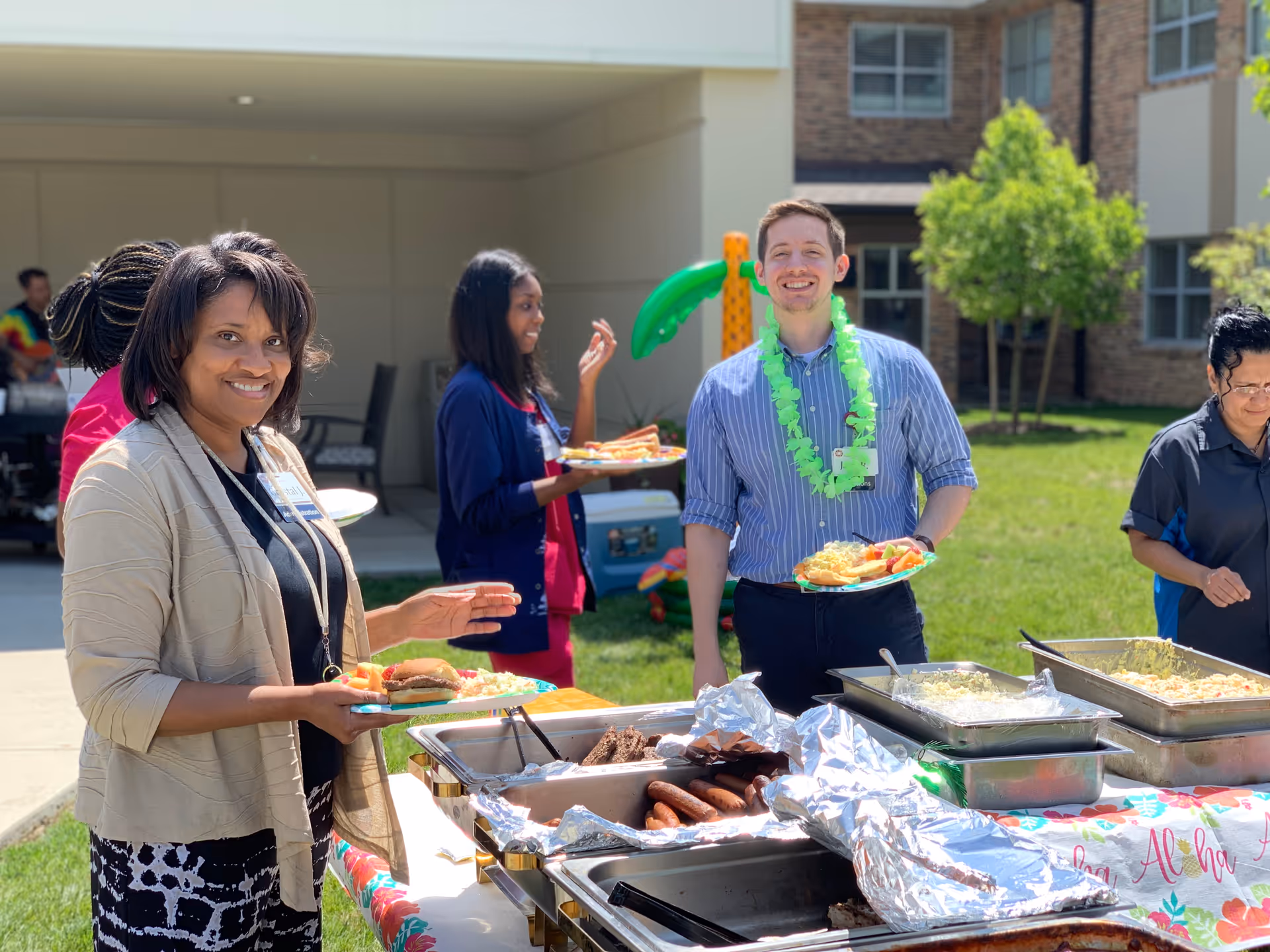 A group of people enjoying an outdoor buffet at a senior living facility. A woman in the foreground holds a plate with a hamburger and sides, smiling at the camera. A man wearing a green lei and a striped shirt stands behind the buffet table with a plate of food, smiling. Other people are serving themselves food from trays on the table. The setting is a grassy courtyard with a building and trees in the background.