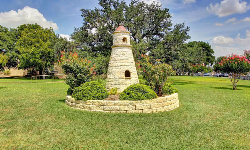 Stone lighthouse-style garden ornament in a circular stone planter surrounded by shrubs on a grassy lawn with trees and a blue sky.